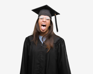Young hispanic woman wearing graduated cap and uniform sticking tongue out happy with funny expression. Emotion concept.