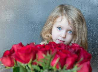 Obraz na płótnie Canvas Charming blonde Caucasian girl looking from behind red roses. The natural raindrops on the window as a background 