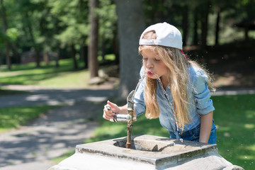 A little cute blonde girl is drinking from Water Fountain in a park. She is wearing a jean t-shirt and a baseball cap