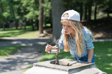 A little cute blonde girl is drinking from Water Fountain in a park. She is wearing a jean t-shirt...