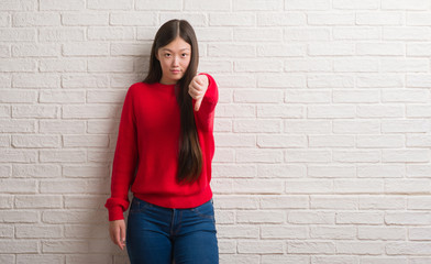Young Chinese woman over brick wall looking unhappy and angry showing rejection and negative with thumbs down gesture. Bad expression.