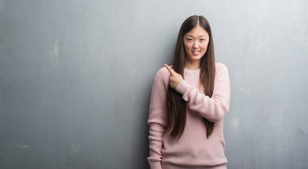 Fototapeta premium Young Chinese woman over grey wall cheerful with a smile of face pointing with hand and finger up to the side with happy and natural expression on face looking at the camera.