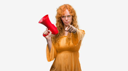 Young redhead woman holding megaphone pointing with finger to the camera and to you, hand sign, positive and confident gesture from the front