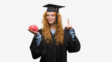 Young redhead student woman wearing graduated uniform holding brain surprised with an idea or question pointing finger with happy face, number one