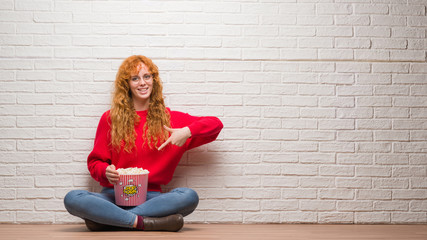 Young redhead woman sitting over brick wall eating popcorn very happy pointing with hand and finger