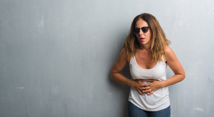 Middle age hispanic woman over grey wall wearing sunglasses with hand on stomach because indigestion, painful illness feeling unwell. Ache concept.