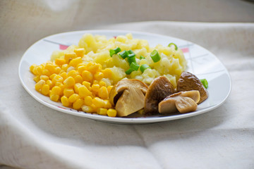 Fried potatoes with cepes and corn in a plate on the table. Fried potatoes with pickled mushrooms and corn.