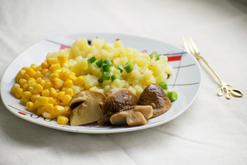 Fried potatoes with cepes and corn in a plate on the table. Fried potatoes with pickled mushrooms and corn.