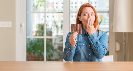 Redhead woman holding chocolate bar at home cover mouth with hand shocked with shame for mistake,...