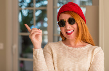 Stylish redhead woman wearing wool cap and sunglasses very happy pointing with hand and finger to...
