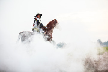 Asian Thai Warrior in traditional armor suit riding horse in white fog with rural farm background. Vintage Retro war costume concept.