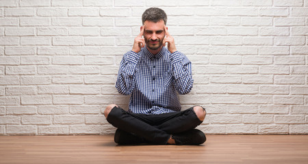 Young adult man sitting over white brick wall with hand on head for pain in head because stress. Suffering migraine.