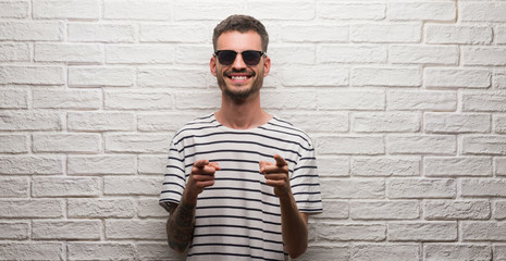 Young adult man wearing sunglasses standing over white brick wall pointing fingers to camera with happy and funny face. Good energy and vibes.