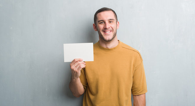 Young caucasian man over grey grunge wall holding blank card with a happy face standing and smiling with a confident smile showing teeth