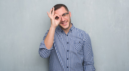 Young caucasian business man over grey grunge wall with happy face smiling doing ok sign with hand on eye looking through fingers