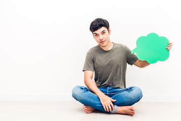 Young man sitting on floor and holding up a speech bubble icon against copy space for adding text with white wall background