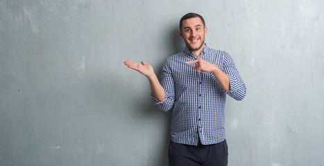 Young caucasian man over grey grunge wall amazed and smiling to the camera while presenting with hand and pointing with finger.