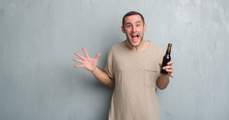 Young caucasian man over grey grunge wall holding bottle beer very happy and excited, winner expression celebrating victory screaming with big smile and raised hands