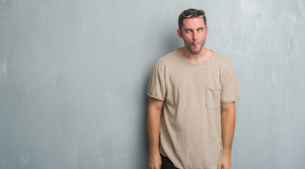 Young caucasian man over grey grunge wall wearing sunglasses making fish face with lips, crazy and comical gesture. Funny expression.