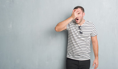 Young caucasian man over grey grunge wall peeking in shock covering face and eyes with hand, looking through fingers with embarrassed expression.