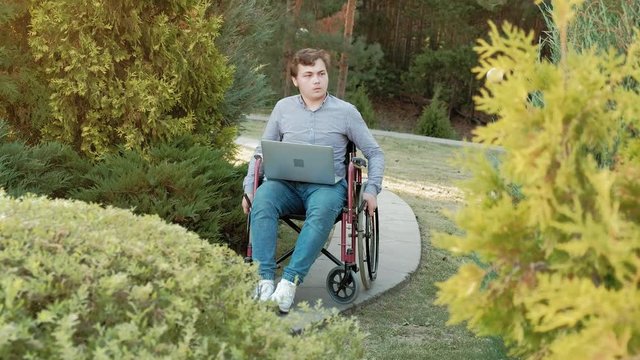A disabled man is sitting in a wheelchair and working on a laptop in the park