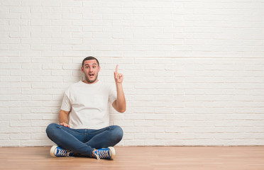 Young caucasian man sitting on the floor over white brick wall pointing finger up with successful idea. Exited and happy. Number one.