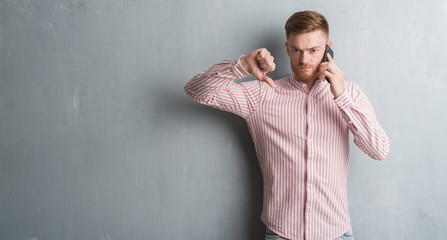 Young redhead man over grey grunge wall talking on the phone with angry face, negative sign showing dislike with thumbs down, rejection concept