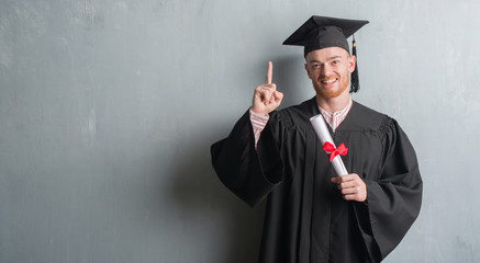 Young redhead man over grey grunge wall wearing graduate uniform holding degree surprised with an idea or question pointing finger with happy face, number one