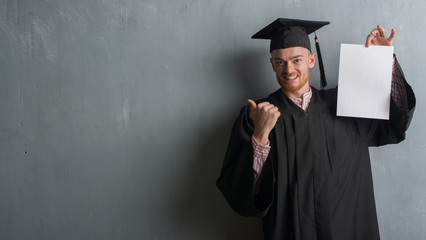 Young redhead man over grey grunge wall wearing graduate uniform holding degree pointing and...
