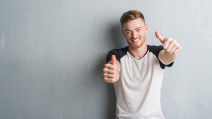 Young redhead man over grey grunge wall approving doing positive gesture with hand, thumbs up...