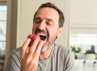 Middle age man eating healthy red apple with a happy face standing and smiling with a confident...