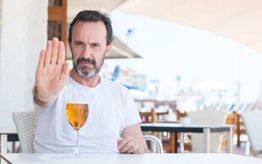 Handsome senior man drinking beer at restaurant with open hand doing stop sign with serious and confident expression, defense gesture