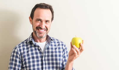 Senior man holding a green apple with a happy face standing and smiling with a confident smile showing teeth