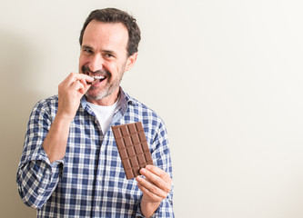 Senior man eating chocolate bar with a happy face standing and smiling with a confident smile showing teeth