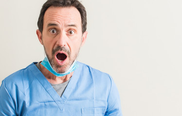 Senior doctor man using mask scared in shock with a surprise face, afraid and excited with fear...