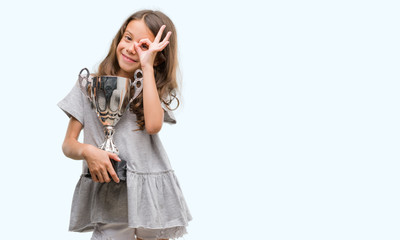 Brunette hispanic girl holding a trophy with happy face smiling doing ok sign with hand on eye looking through fingers