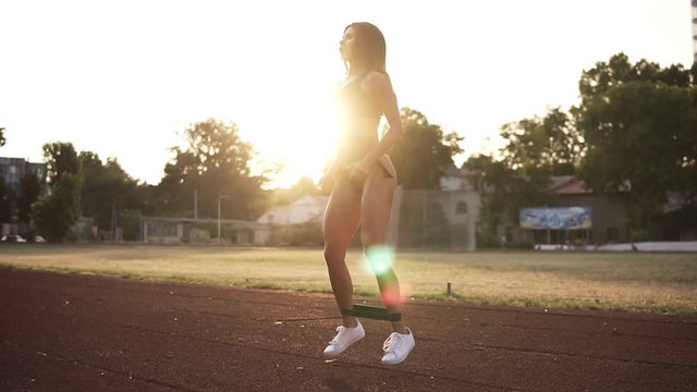 Young woman athlete exersices with resistance band outdoors at sunset or sunrise. Attractive woman wearing black bikini and white sheakers