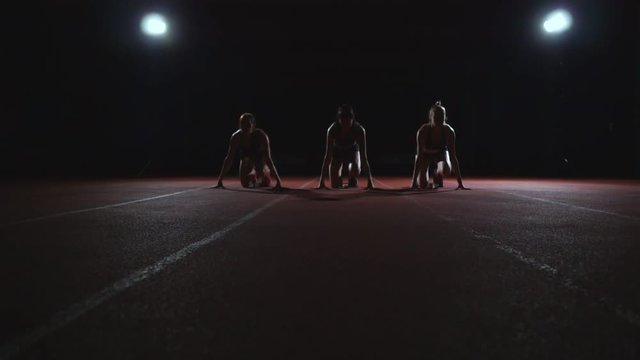 Three girls in black clothes are in the starting pads to start the race in the competition in the light of the lights and run towards the finish