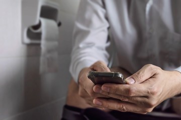 Man sitting on toilet bowl while playing mobile phone - health problem concept
