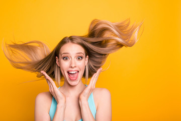 Portrait of cute straight-haired blonde caucasian smiling girl, wearing casual blue shirt, amazed, showing excitement, wind blows hair. Isolated over yellow background