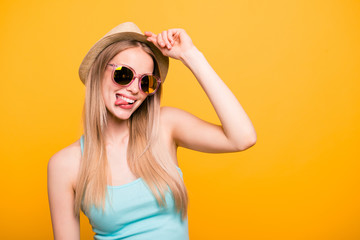 Attractive nice cute straight-haired blonde caucasian smiling girl, wearing casual shirt, cap, sun glasses. Enjoying trip. Grimacing, showing tongue out. Isolated over yellow background
