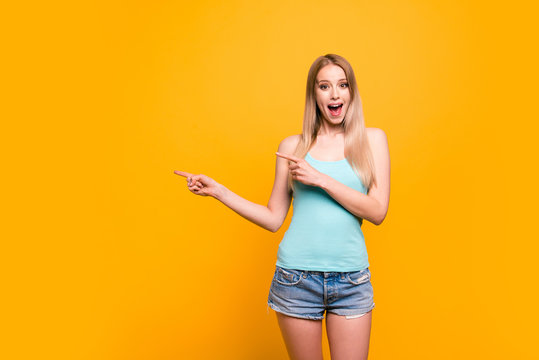 Attractive cute straight-haired blonde caucasian smiling girl, wearing casual blue shirt and jeans shorts, amazed, pointing showing with two fingers to object. Isolated over yellow background