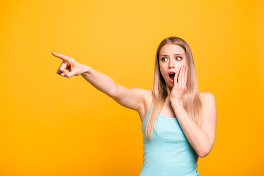 Attractive cute straight-haired blonde caucasian shocked girl, wearing casual blue shirt, pointing showing far way with finger. Isolated over yellow background