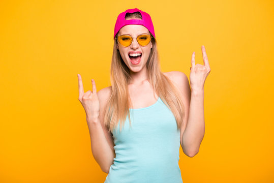 Attractive cute straight-haired blonde caucasian funny girl, wearing casual blue shirt and vivid bright pink cap, showing double horns sign with hands. Isolated over yellow background