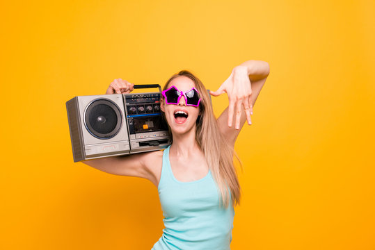 Attractive nice cute straight-haired beautiful blonde caucasian smiling girl, wearing casual shirt, sun glasses star shape. Holding boom box, enjoying music. Isolated over yellow background