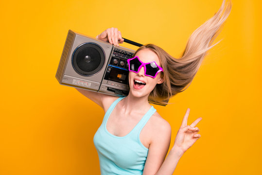 Portrait of cool young blond lady, wearing summer glasses showing v-sign holds a tape recorder on her shoulder isolated on bright yellow background.