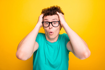 Young attractive handsome shocked guy wearing casual green polo t-shirt and glasses, holding his head with hands showing surprising. Isolated over vivid shine bright yellow background