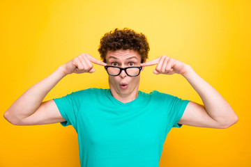 Handsome curly-haired shocked young guy wearing casual green t-shirt, showing surprised gesture, putting glasses down. Isolated over yellow background