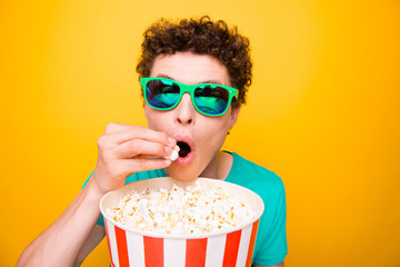 Young attractive handsome funky guy wearing casual green polo t-shirt and 3d sun glasses, eating pop corn, watching cinema. Isolated over vivid shine bright yellow background © deagreez