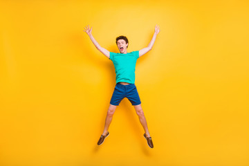 Fototapeta na wymiar Full size length body picture of handsome curly-haired funny young guy wearing casual green t-shirt, shorts, shoes, jumping in air, hands up, star figure. Isolated over yellow background